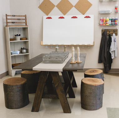 image of a classroom with natural workspace, hanging paint shirts, and shelves of interesting art materials