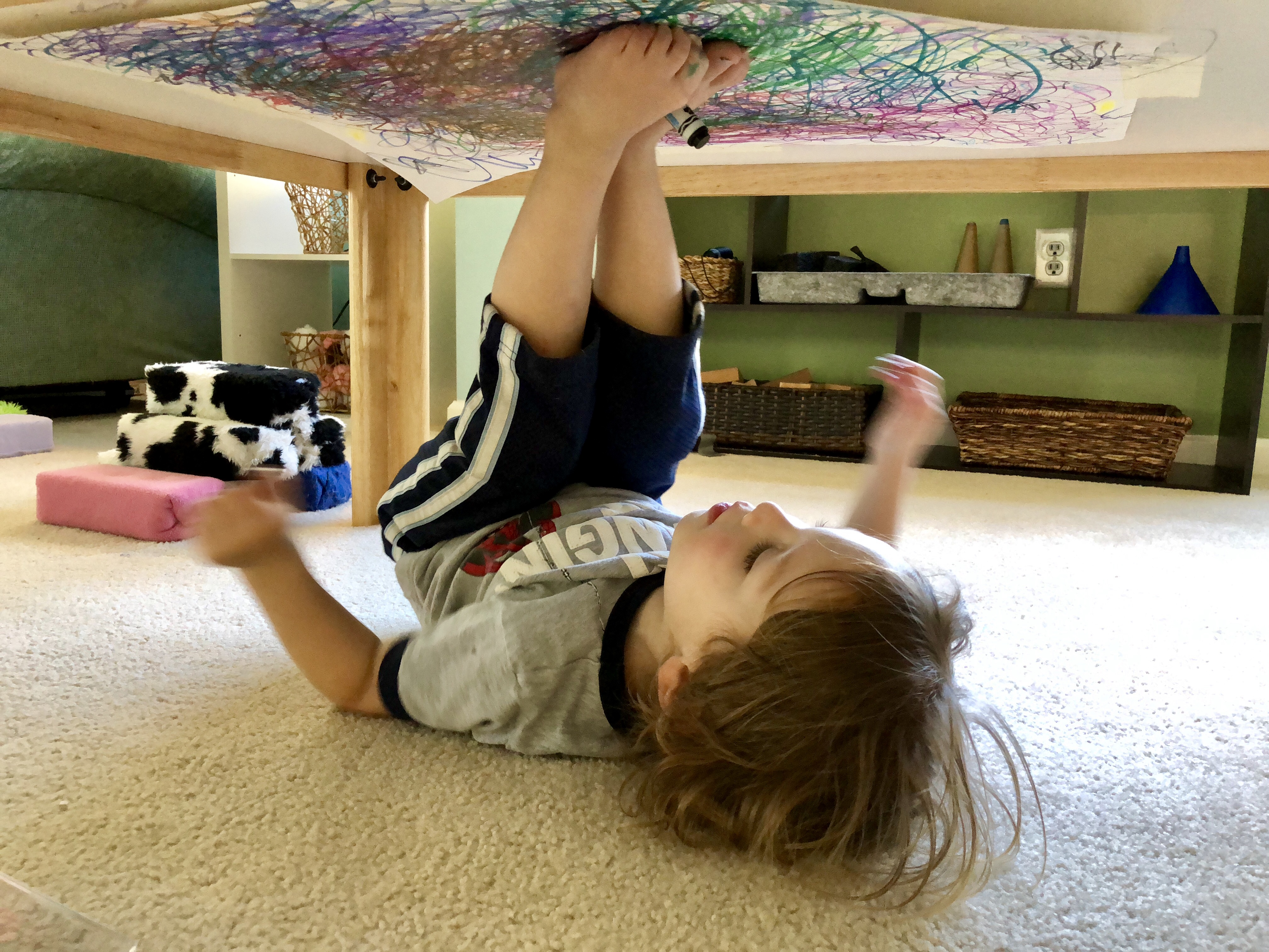 boy holding a marker in between his feet and coloring on the underside of a table