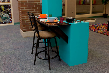 two stool pulled up to a large play kitchen. Plates and cups are on a small table mounted to the kitchen oven.